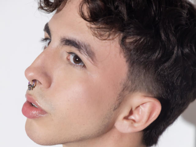 BrahmBelial tem um piercing no nariz, lábios rosados e brilhantes, e cabelos cacheados e castanho-escuros. Sua pele é clara, e ele está olhando para o lado com olhos castanhos..
