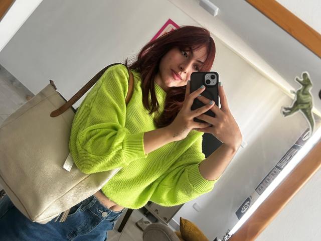 SpencerLe, con cabello rojo, vistiendo un suéter verde neón, jeans azules y un bolso beige, tomando una selfie con un teléfono negro, mostrando una cara linda..