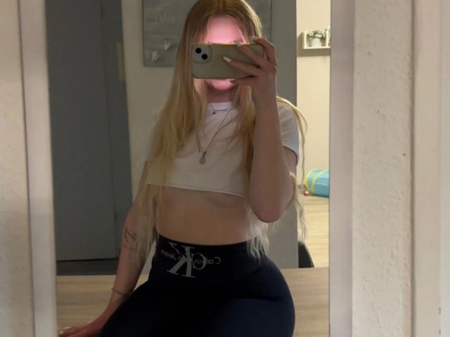 RoyalBlondie-hot with long blonde hair, white crop top, exposing small tits, black high-waisted pants, holding phone in mirror selfie, pink face blur, tattoo on left arm.