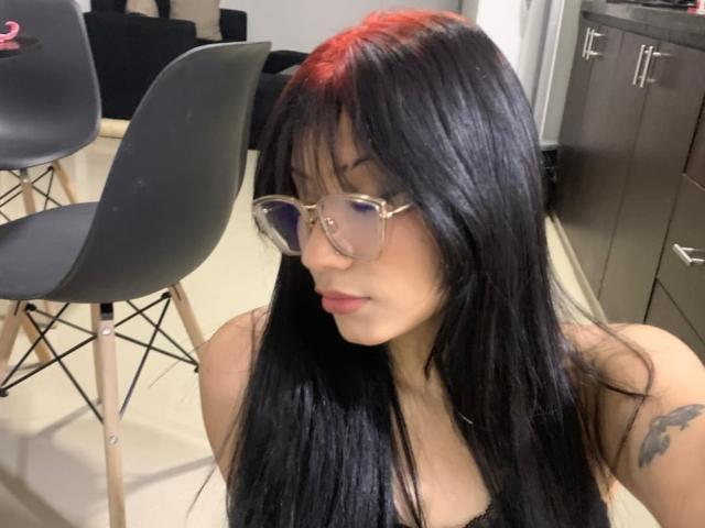 NaomiOsaka with long black hair, red highlights, and glasses, showing her tits in a black top, has a tattoo on her right arm, sitting near a chair.