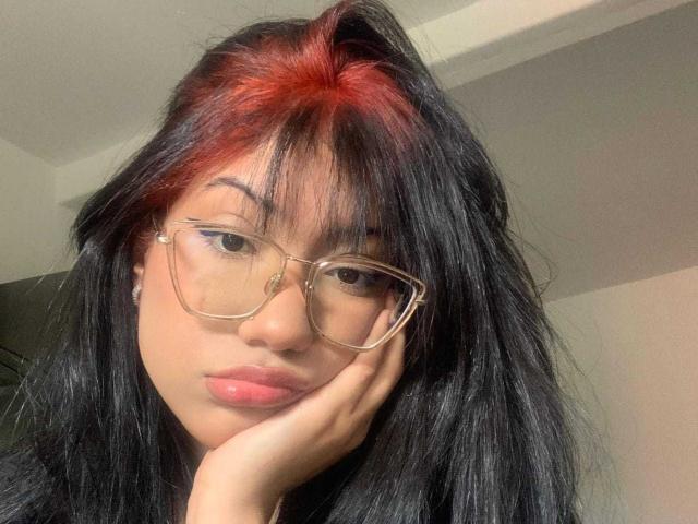 NaomiOsaka has black hair with red highlights, wearing glasses, and resting her face on her hand, with pink lips and a neutral expression.