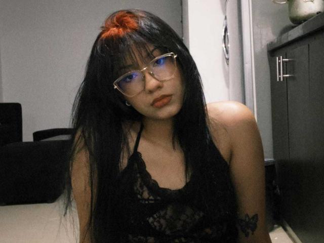 NaomiOsaka with long black hair and red top knot, wearing glasses and black lace top, showing tattoos on her right arm, sitting in a dimly lit room.