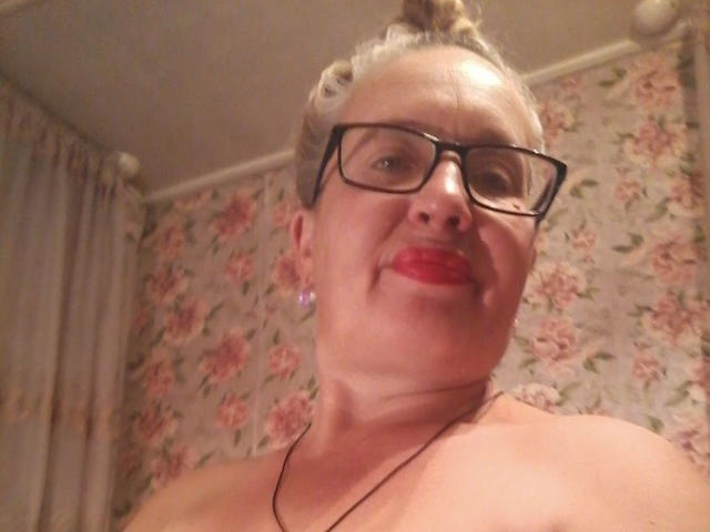CerdAyaz has blonde hair in a bun, black glasses, red lipstick, and is topless with visible shoulders, wearing a black necklace, and smiling.