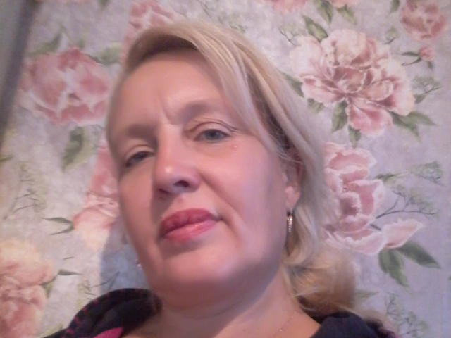 CerdAyaz has pale skin, blonde hair, red lipstick, small earrings, and a slight smile. Her tits are not visible. She's wearing a black top with pink accents.