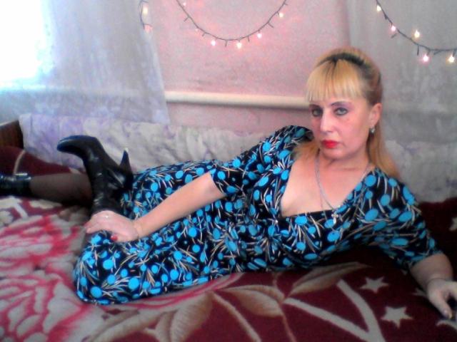CerdAyaz with blonde hair, blue and black patterned dress, black boots, laying on red floral blanket, looking at camera, red lipstick, white walls, string lights in background.