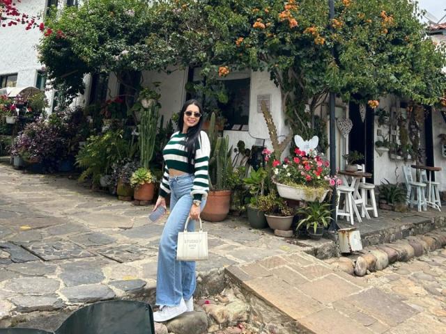 IsabellaJames with long black hair, wearing a green and white striped sweater, blue high-waisted jeans, white sneakers, holding a white purse, standing on a stone sidewalk.
