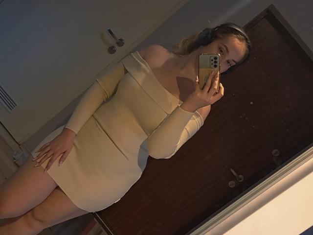 AliceNewX in a tight, off-shoulder beige dress, holding a phone, covering her face, with headphones on, standing in front of a wooden door, showing her ass.