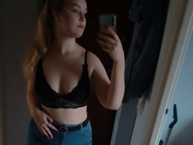 AliceNewX, blonde, wearing black lace bra, showing her tits, blue jeans, mirror selfie, holding phone, dim room, casual, confident, slightly blurred, hand on hip.