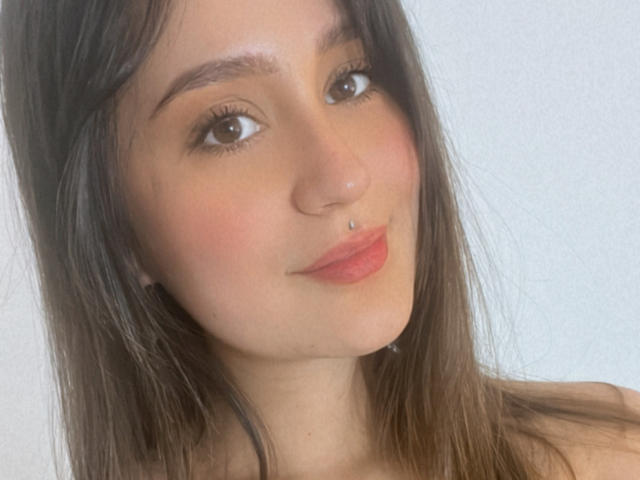 NinaBryons has a cute face, brown eyes, and a silver nose piercing. She's smiling with pink lipstick and has long brown hair. Boobs aren't visible in this shot.