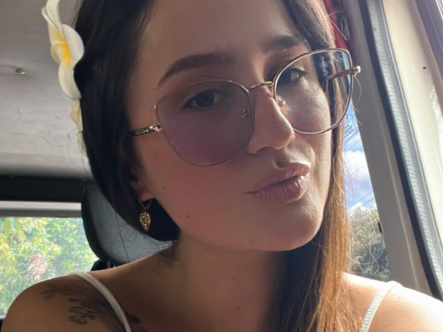 NinaBryons has brown hair, a white flower in her hair, pink-tinted aviator sunglasses, and a cute pout. She's wearing a white top with black earrings showing.