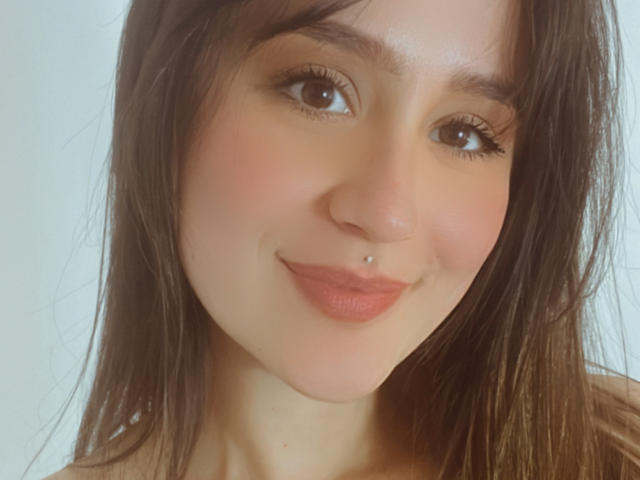 NinaBryons has a cute face with brown eyes, long brown hair, a nose piercing, and pink lips. Her skin is fair and she's smiling softly.