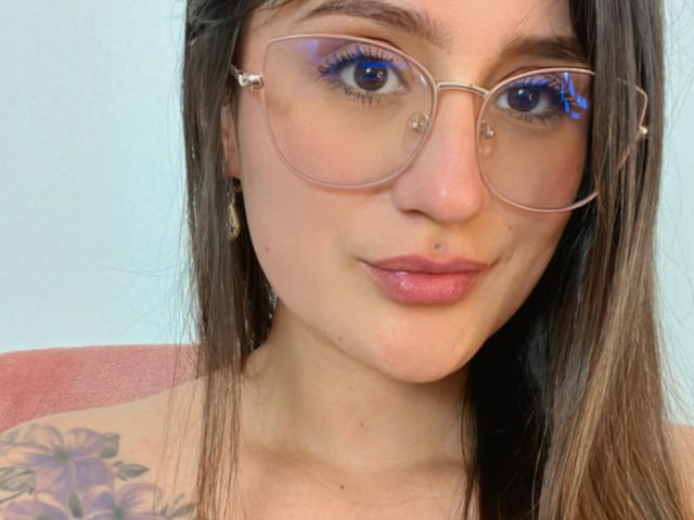 NinaBryons with glasses, blue eyeliner, and nose ring, showing a slight smile, has a tattoo on her shoulder, and has a lip piercing. Her tits are not visible.