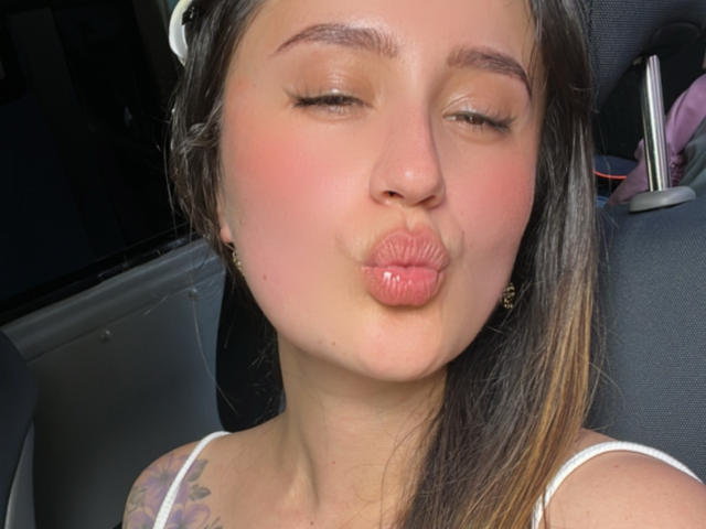 NinaBryons has her eyes closed, puffy pink lips puckered in a kiss, pink blush on her cheeks, brown hair, white tank top, and a tattoo on her left shoulder.