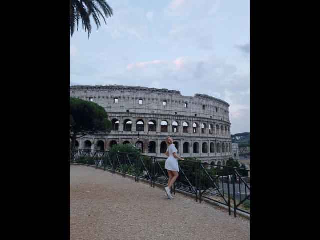 CatIlusionxx-hot stands in front of the Colosseum, wearing a white dress and white sneakers, with a gravel path and green fence in the foreground, palm tree on the left.