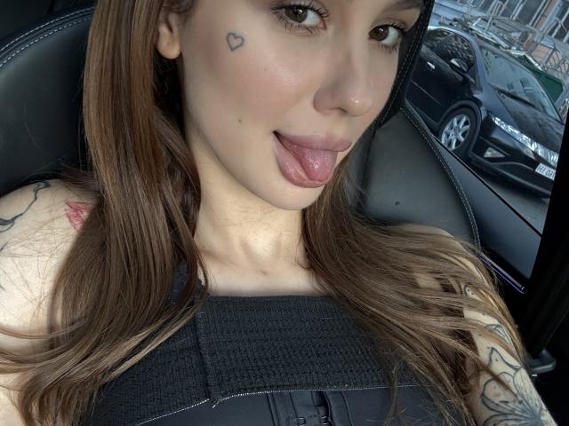 SensualMila with brown hair, tongue out, small heart tattoo under right eye, black seatbelt, tattooed arm, and visible car seat in background. Boobs, tits, and pussy not visible.