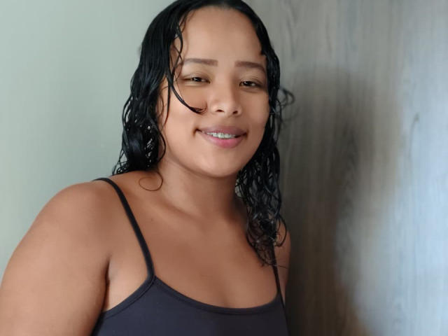 VelvetAndNoiir has wet black hair, brown eyes, and a cute smile. She's wearing a black spaghetti-strap top, showing her small tits and light brown skin.