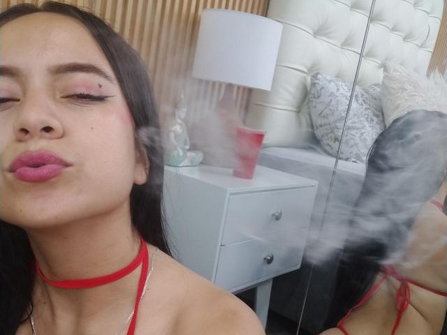 JessyLenon with pink lipstick, red choker, and smoky breath in selfie. Reflective mirror, white bed with textured pillows, and red cup in background. Her eyes are half-closed.