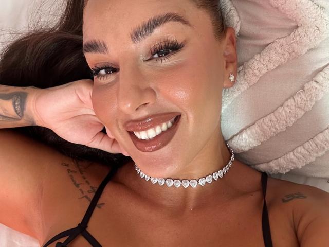 CheyenneSinLimites smiling with glossy brown lips, wearing a diamond choker, black strap top, and showing tattoos on her arm and shoulder. Her brown eyes are slightly winking.
