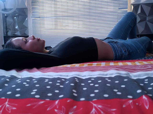 TinyLaurent is lying on a bed, wearing a black top and blue jeans, with her ass raised and her head on a pillow, looking to the side.
