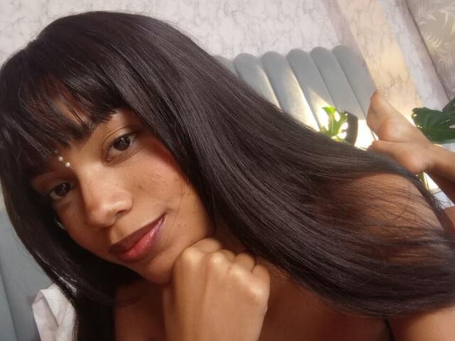 AylaCallowey with long black hair, brown eyes, and a small white dot on her forehead, resting her chin on her hand, showing a subtle smile and bare shoulders.