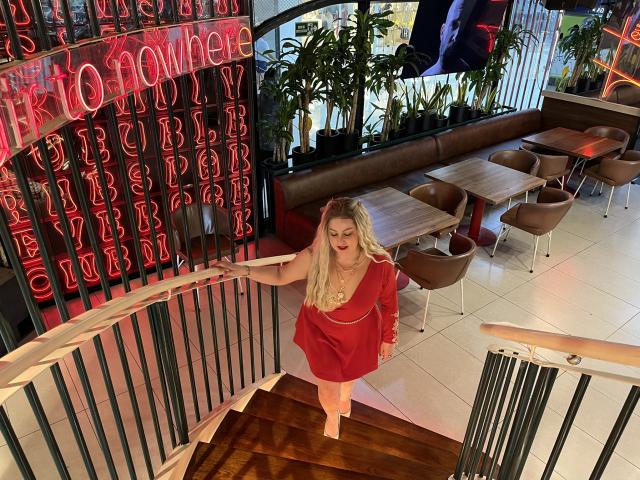 SamanthaGold with blonde hair and red dress, holding railing, looking up, brown wooden tables, brown chairs, red neon lights, and potted plants in a modern cafe.