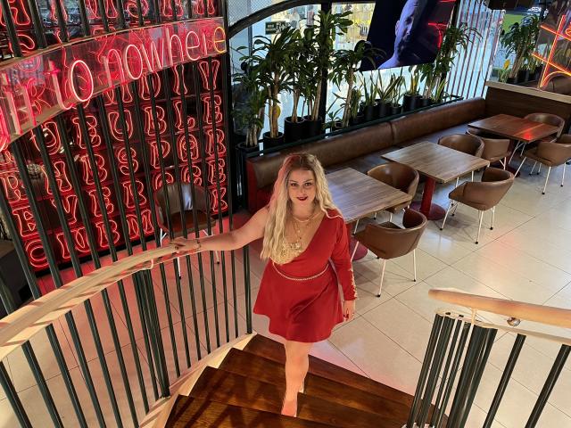 SamanthaGold with long blonde hair, red dress, and red lipstick, standing on stairs, hand on railing, looking up, in a modern restaurant with brown chairs and neon signs.