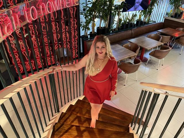 SamanthaGold with long blonde hair, red dress, and gold necklace, standing on wooden stairs, holding railing, wearing red lipstick, looking down at camera, in a brightly lit room.