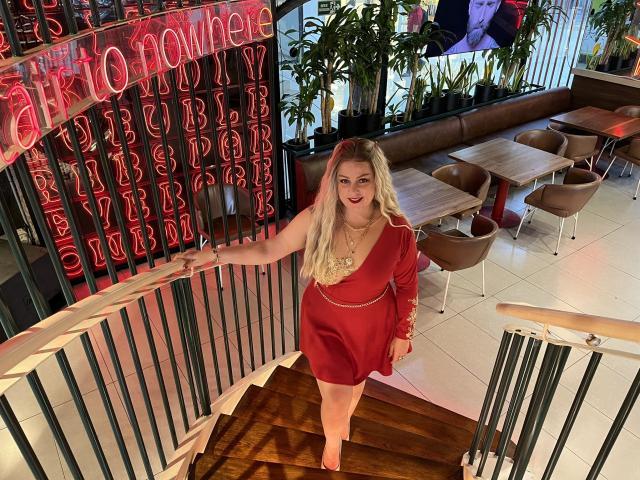 SamanthaGold with blonde hair, red dress, and red heels, standing on wooden stairs, smiling, holding railing, showing her ass, in a modern cafe with neon lights.