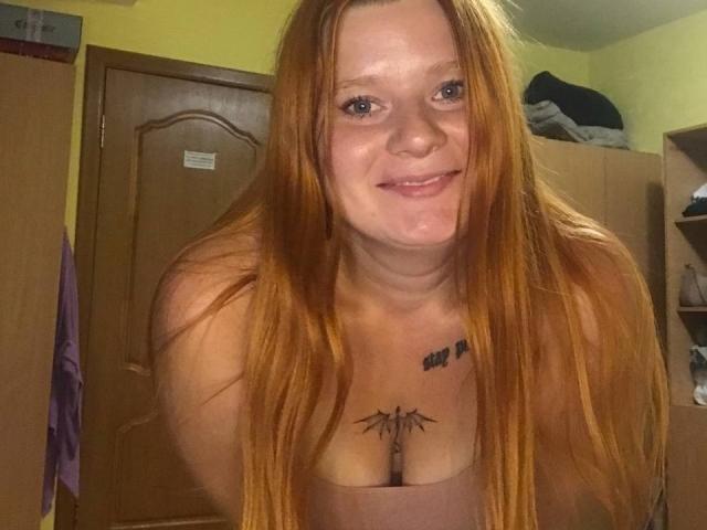 PollyKatie with long red hair, smiling, showing off her tits and tattoos, wearing a pink tank top, leaning forward in a bedroom.