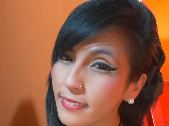 DaniaHot69 ha capelli neri lucidi, un eyeliner nero marcato, rossetto rosa, orecchini di perle e un piccolo piercing al naso, e sorride con un leggero movimento del capo..