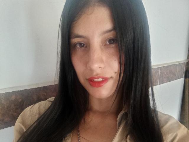 MadissonFontaine has long black hair, brown eyes, red lipstick, and a beauty mark on her right cheek. She's wearing a beige shirt and a silver necklace.