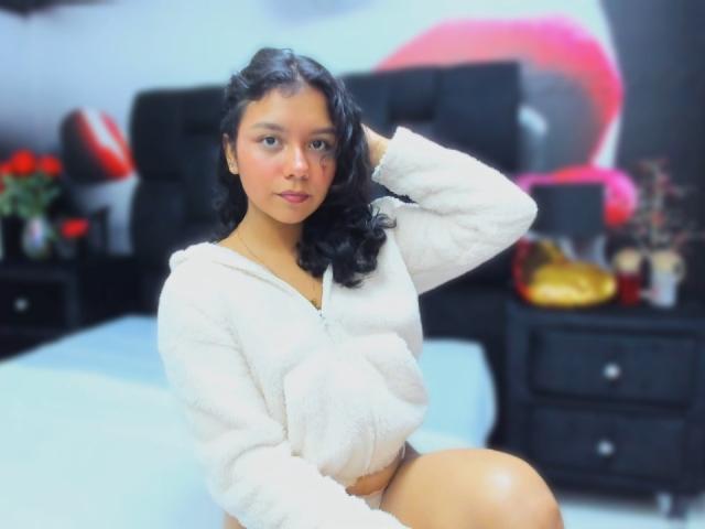 JuliaBennett with dark curly hair, wearing a white fluffy hoodie, sitting on a bed, hand in hair, showing a cute face, her pussy barely covered by the hoodie.