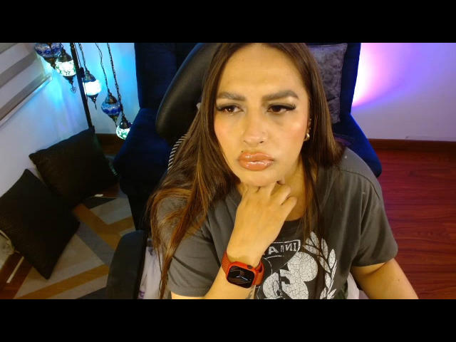 EmiliaMontserrat has long brown hair, a nose ring, and is wearing a gray t-shirt with a black chair behind her. She's pouting with her finger on her chin.
