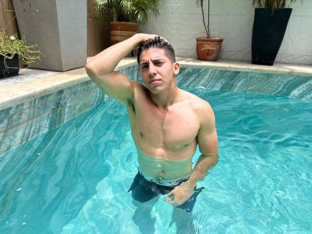 SwingerTeam, shirtless, muscular, standing in a blue-tiled pool, water up to his thighs, hand on head, looking annoyed, dark wet hair, black swim trunks, potted plants in background.