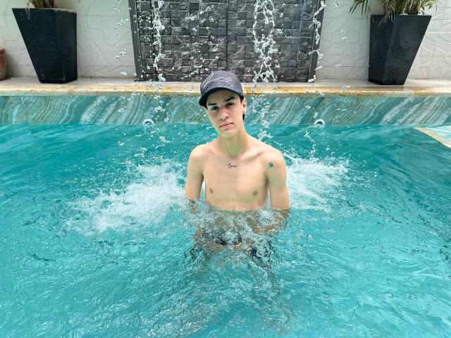 Shirtless SwingerTeam with a small tattoo on the right shoulder, standing in a pool, water splashing around, wearing a dark cap, visible blue swim trunks, and a slight smile.