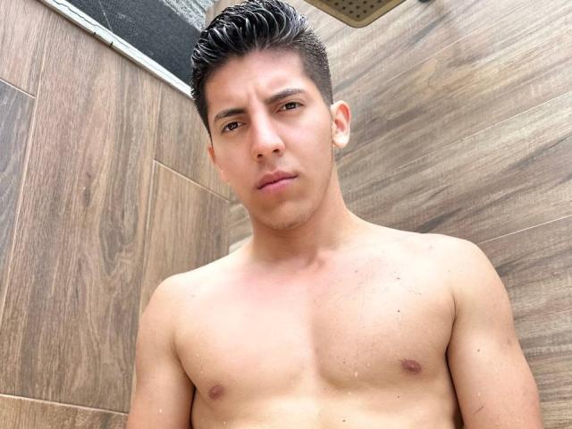 Shirtless SwingerTeam with wet, slicked-back black hair, light brown skin, and a serious expression, standing in a wooden-tiled shower. Small, slightly raised nipples visible.