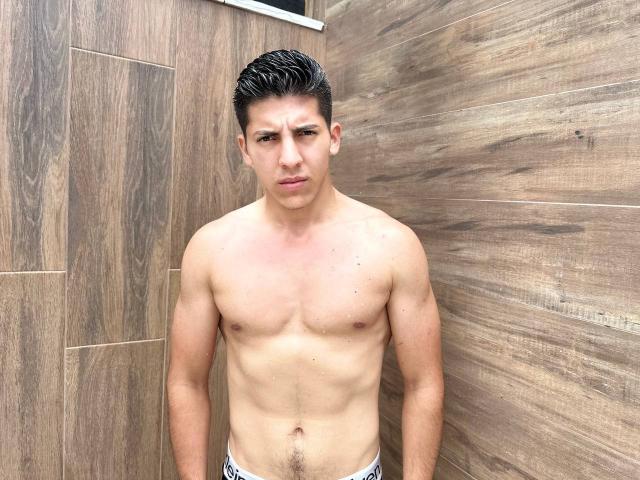 Shirtless SwingerTeam with a pained expression, standing in a wooden-walled room. He's got a muscular, toned body, short dark hair, and white underwear with a black pattern.