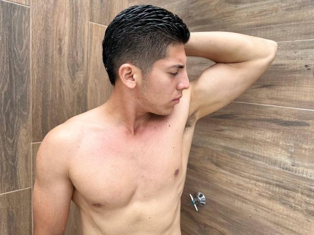 Shirtless SwingerTeam with wet black hair, raised arm, showing armpit hair, and muscular, tan, slightly red chest. Wooden wall background. Water droplets on skin.