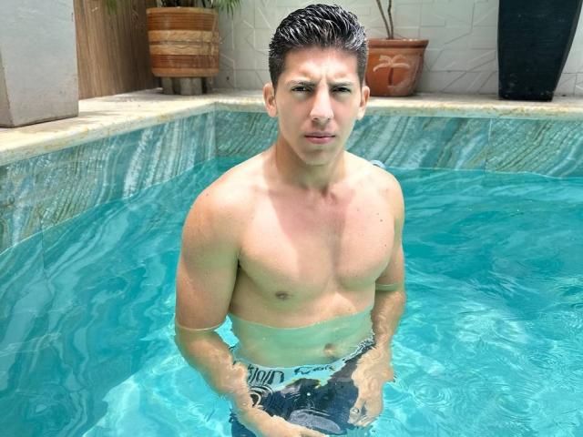 Shirtless SwingerTeam with wet, dark hair stands in a blue-tiled hot tub, water up to his waist, wearing black and white swim trunks, hands near his cock.