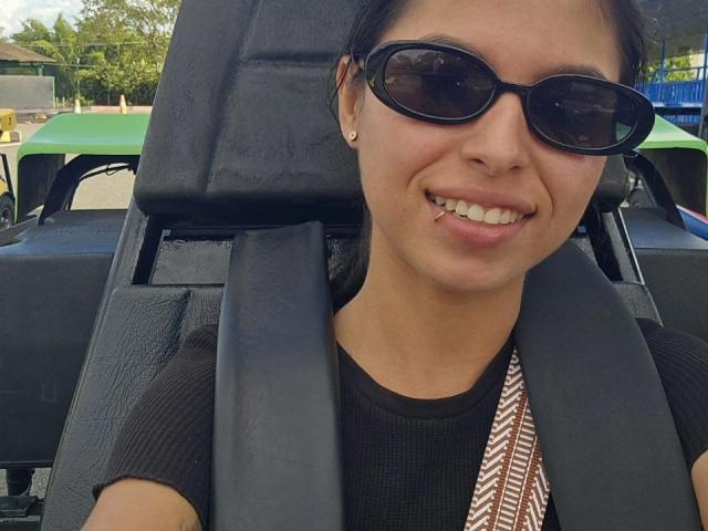 JuliaLanne with black sunglasses, smiling, has a toothpick in her mouth, wearing a black shirt and seatbelt, brown patterned strap, and black ear studs, in a green vehicle.