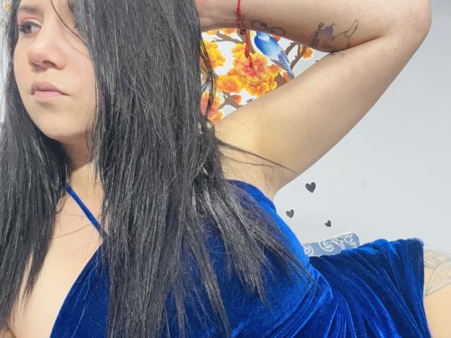 Kkatalina with long black hair, blue velvet dress, arms raised, tattoo on arm, subtle makeup, red bracelet, looking side, visible cleavage, small black hearts on right.