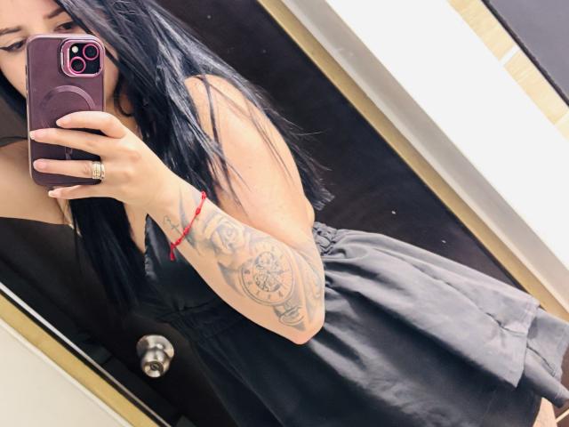 Kkatalina with long black hair, wearing a black dress, holding a purple phone, showing a tattooed arm with a red bracelet, in a mirror selfie.