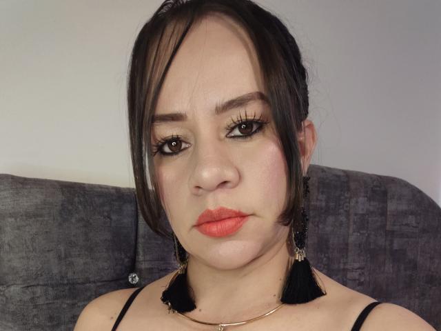 CharllotI has medium skin, black hair in a side ponytail, thick black eyeliner, orange lipstick, black tassel earrings, and a gold necklace, sitting on a gray couch.