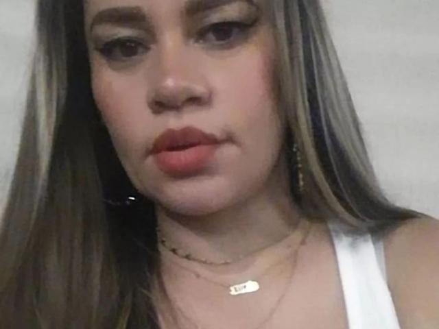 CharllotI with pouty pink lips, long brown hair, and a white tank top, wearing a gold necklace, looking at the camera with slightly parted pink lips.