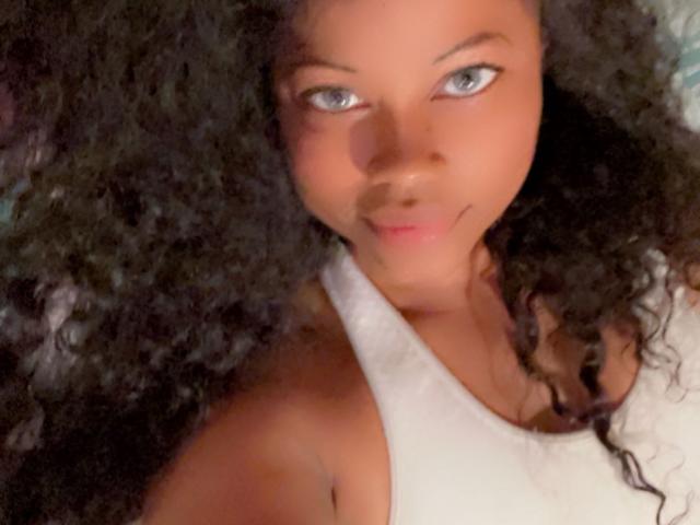 Cincia69 has curly black hair, light brown skin, blue eyes, and is wearing a white tank top, smiling slightly at the camera with a relaxed, confident expression.