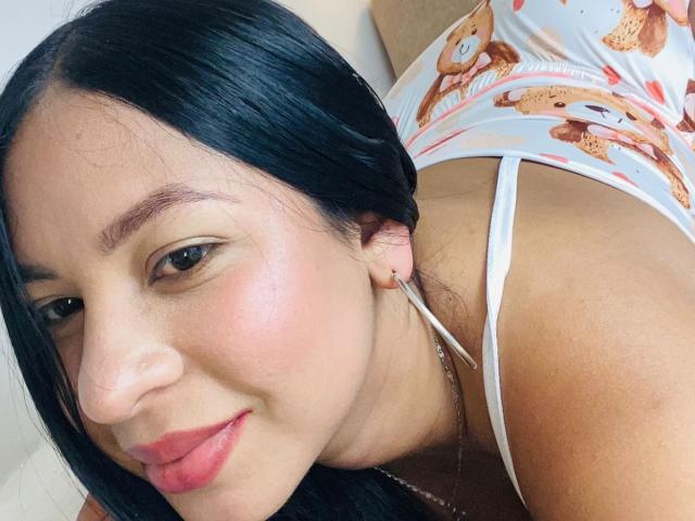 SofiaaTailor with black hair, red lips, and a cute smile, wearing a white teddy bear print top, showing her small tits and bare ass. She's leaning forward.
