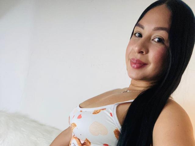 SofiaaTailor with long black hair, pink lipstick, and a white dress with orange and brown patterns, showing her small tits and smiling at the camera.