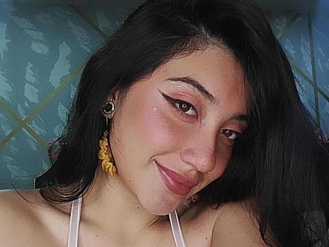 ArianaZahar has black hair, pink eyeshadow, and a small zit on her cheek. She's smiling with yellow earrings and white straps visible on her chest.