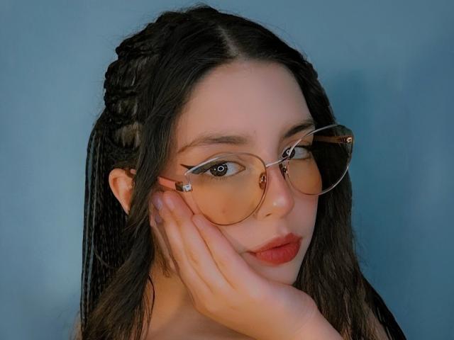 PaolaLyon with braided hair, wearing tortoiseshell glasses, has her hand on her cheek, showing her red lipstick and brown eyes. Her ass is not visible.