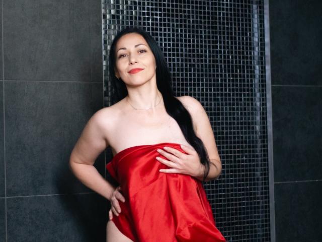 LiluGrants with long black hair, red lipstick, and pale skin, standing nude, covering her tits with a red satin cloth, hand on hip, smiling, in a dark tiled shower.